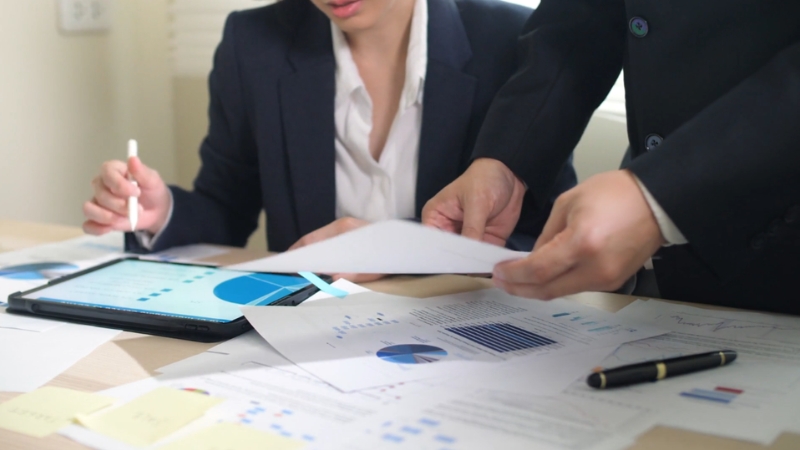 Two professionals review financial charts and reports on a desk with documents, a tablet, and a pen