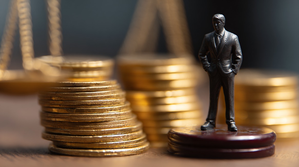 Stacks of gold coins with a figurine of a businessman and scales of justice in the background