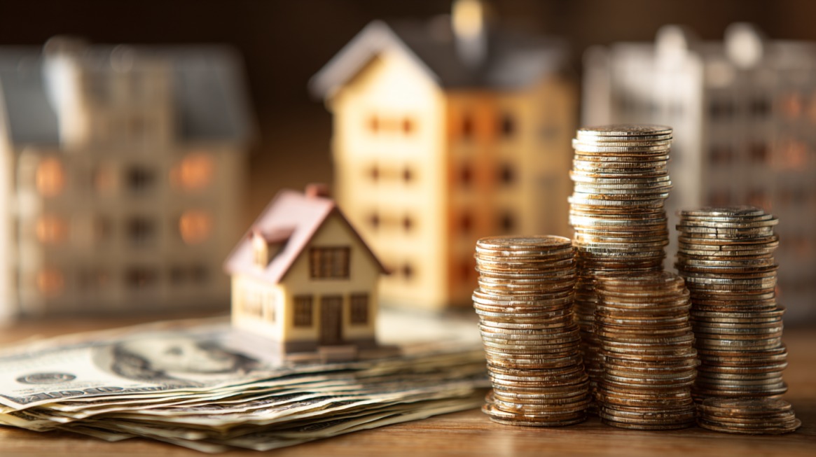 Stacks of coins and small model houses placed on a table with dollar bills underneath