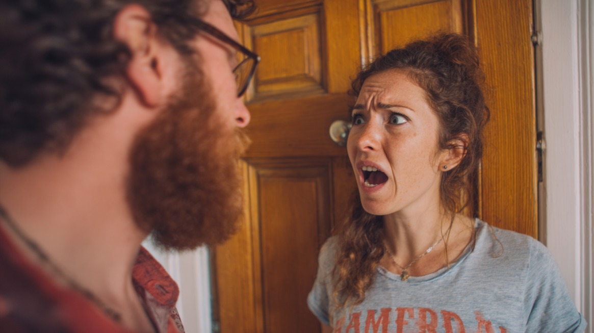A woman standing in front of a wooden door looking upset and yelling at a man facing her