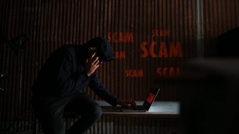 A man in a hoodie uses a phone and laptop to carry out a scam