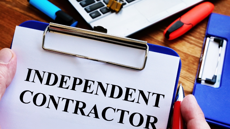 A clipboard with the words Independent Contractor held over a desk with office supplies and a laptop