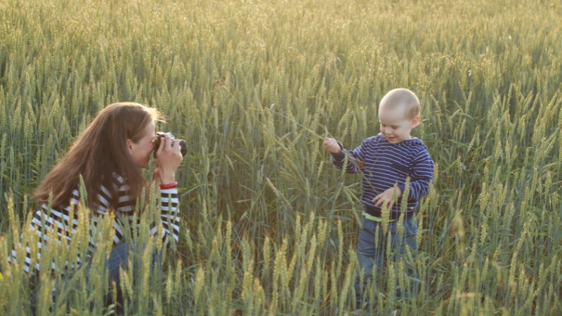 A woman takes a photo of a smiling child standing in a field at sunset