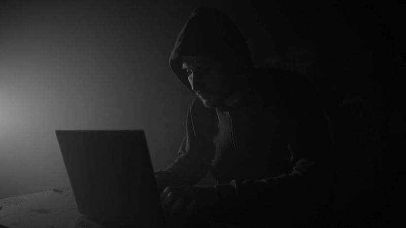 Hooded individual working on a laptop in a dark room, suggesting insider cyber theft of confidential data