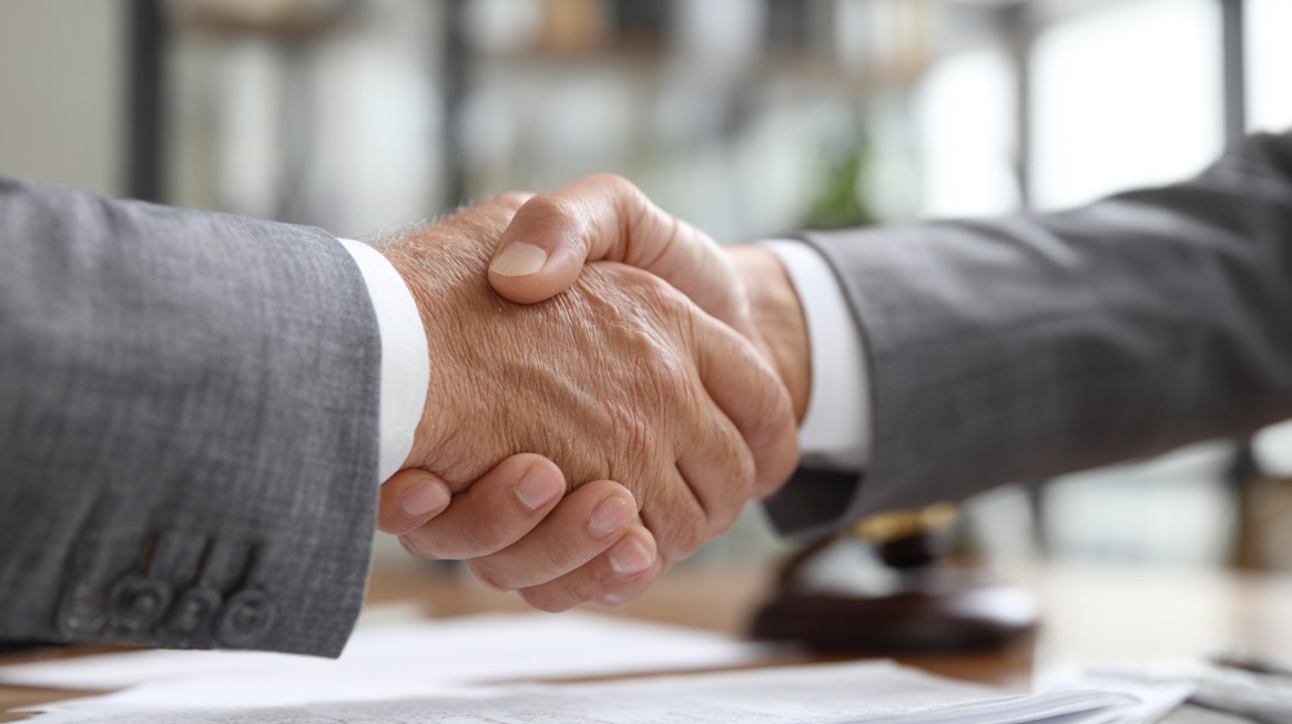 Two people in business suits shaking hands across a desk with legal documents
