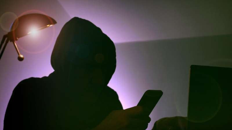 Person in a hoodie checking a phone near a laptop, reflecting unauthorized access to online accounts