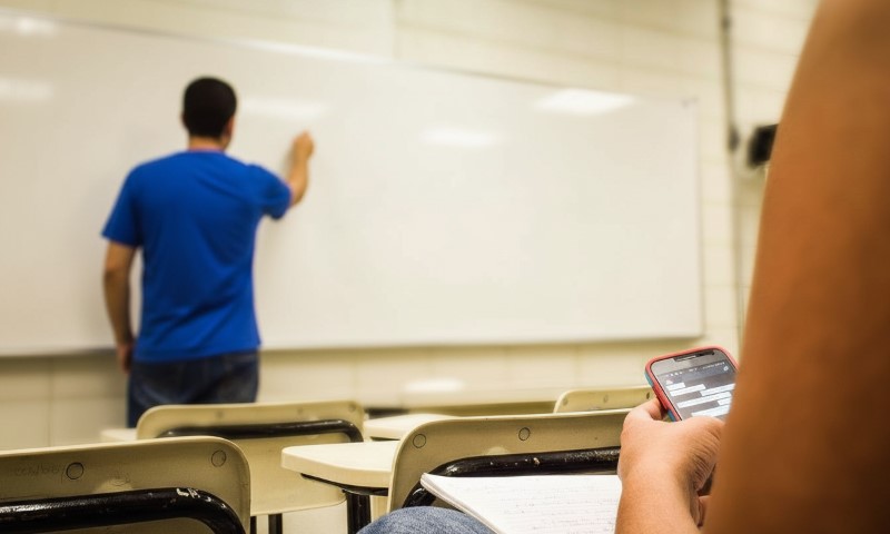 A person in a blue shirt writes on a whiteboard in a classroom, while another person in the foreground uses a smartphone