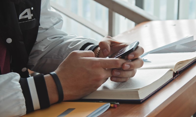 A person in a jacket, seated at a desk with an open book and notebooks, is focused on a smartphone
