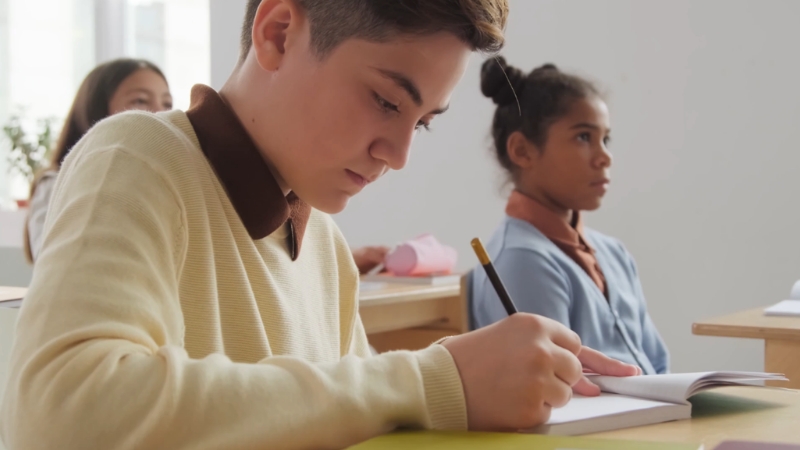 Student writing at a desk in a classroom with another student seated behind