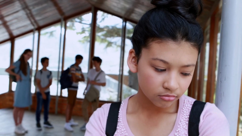 Student standing alone in a school hallway while other students talk in the background